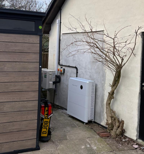 - Flux Solar Energy White outdoor appliance (likely heat pump) on a concrete slab beside a bare-tree and a pale-wall building, with a wooden shed on the left.