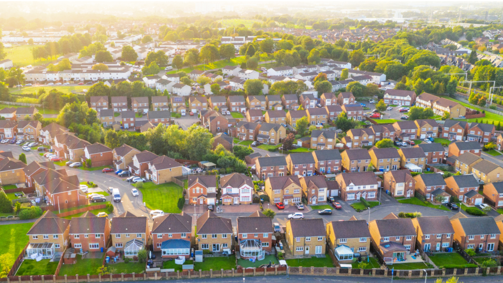 Aerial view of a suburban housing estate with rows of brick houses, green lawns, and tree-lined streets at sunset.